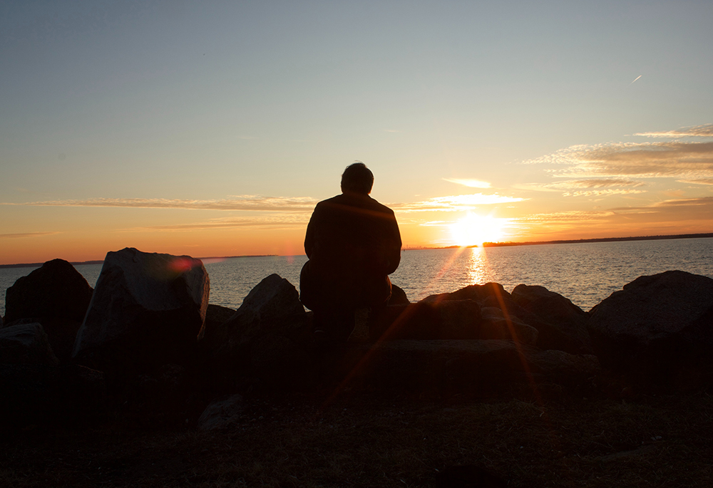 A silhouette of a man sitting on a rock during sunset is shown. (Unsplash/Dylan)