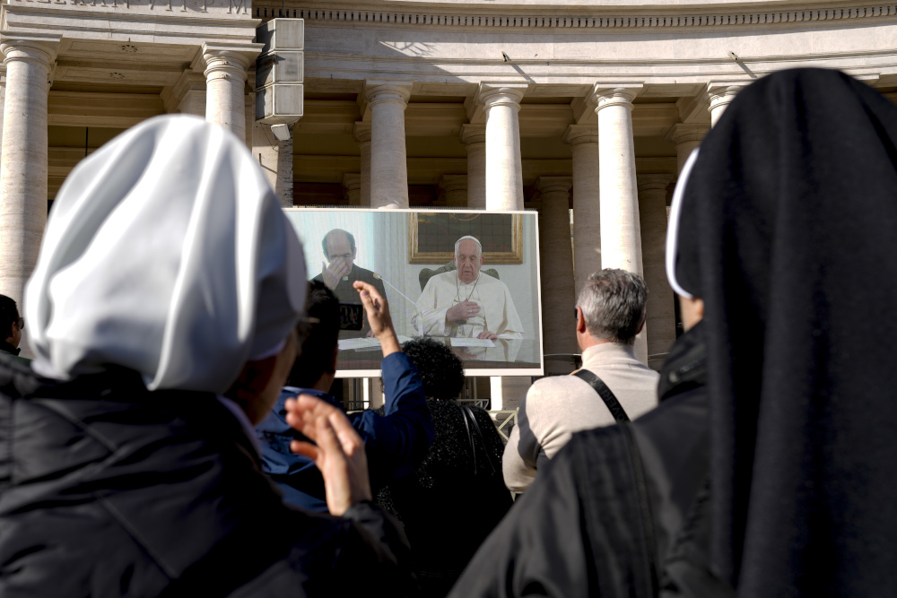 Pope Francis and a priest can be seen on a screen in front of a crowd with the backs of the heads of two religious sisters immediately in front of the camera