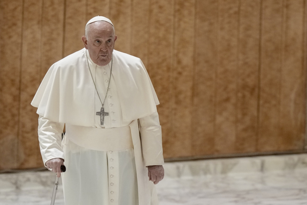 Pope Francis walks with a serious expression with a cane in front of a wooden wall and marble floor