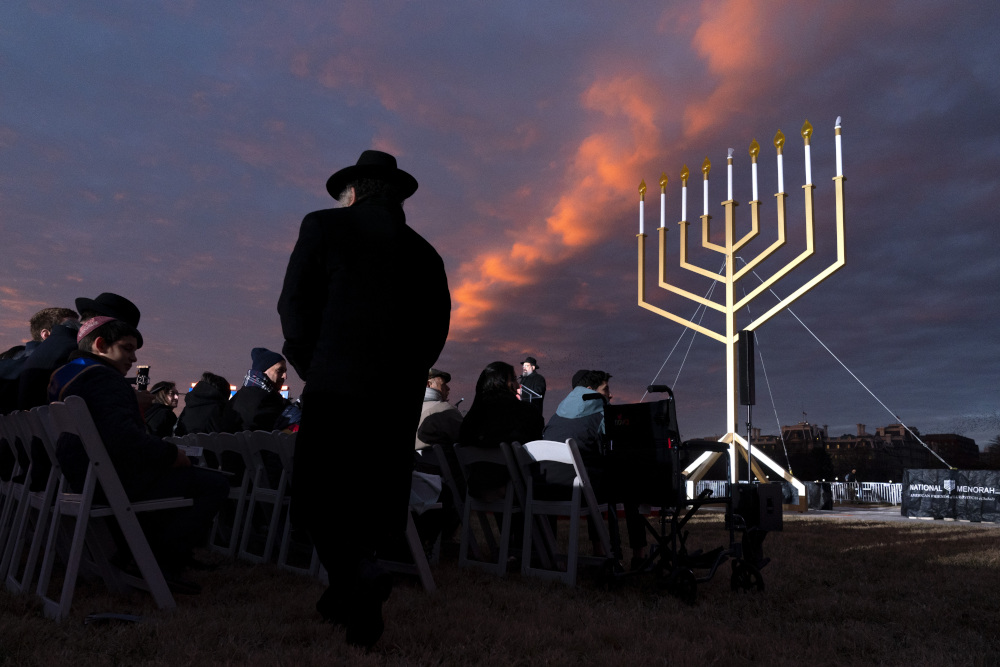 A very large menorah stands in the darkening sky with orange clouds before a crowd