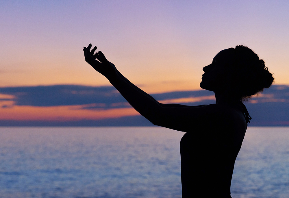 A woman raises her arms against a background that includes a body of water and a sunset. (Unsplash/William Farlow)