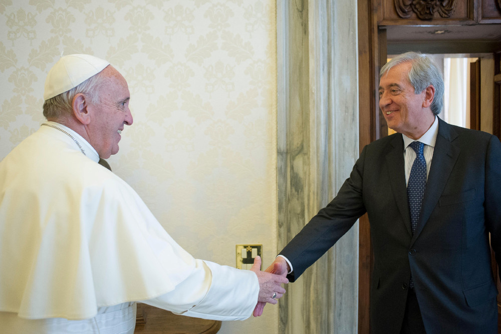 An older white man wearing a suit shakes Pope Francis' hand in a room with white patterned wallpaper