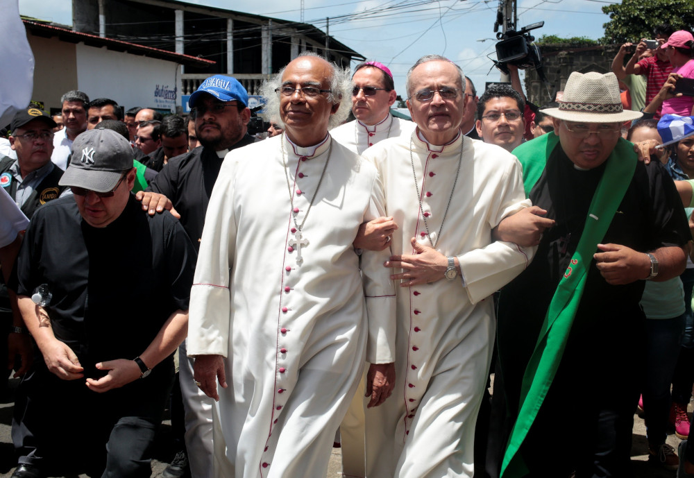 Three men in white cassocks stand among laymen and priests. Two of the men in white cassocks link arms.