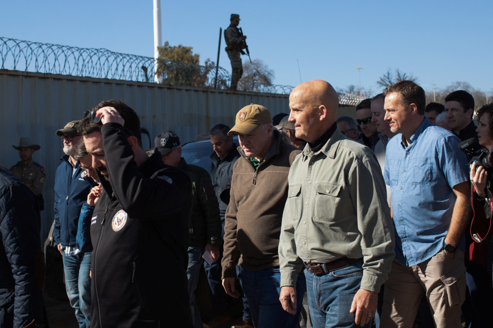 A group of white men in casual clothes walk along a concrete wall topped with barbed wire