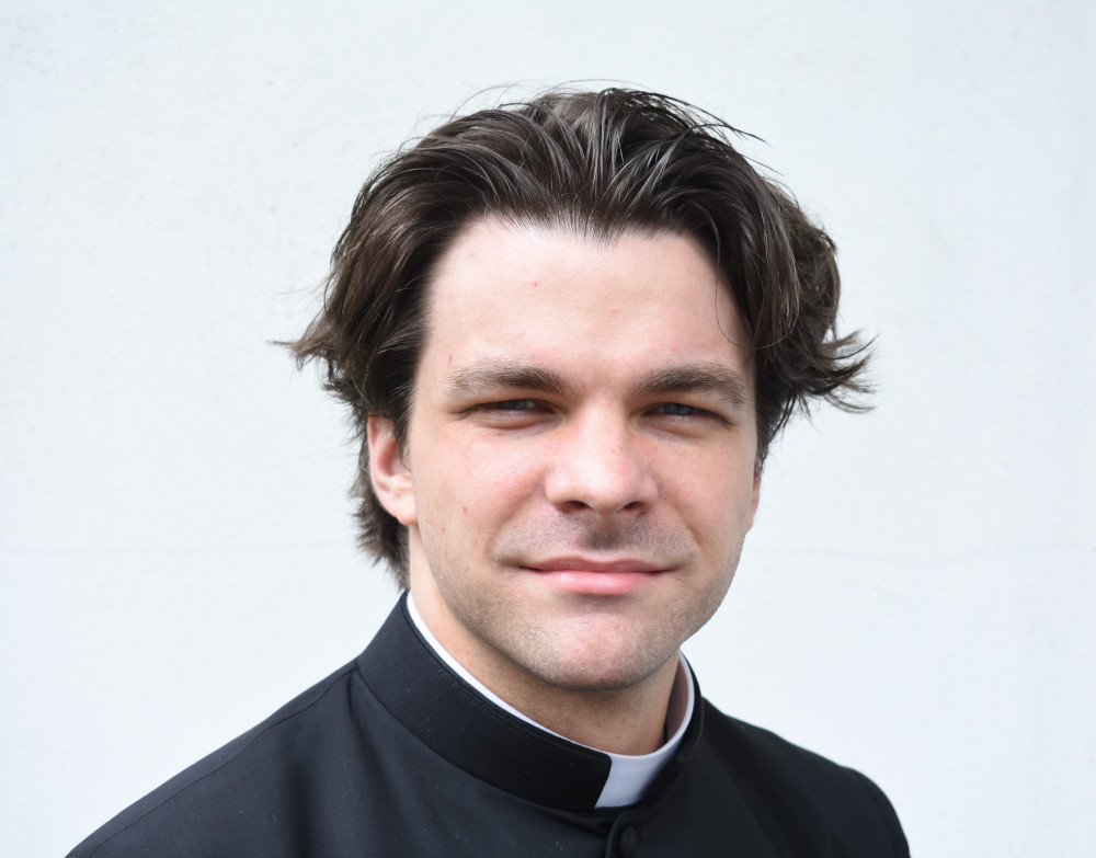 A white man with a swoopy brown haircut wears a clerical collar and stares at the camera