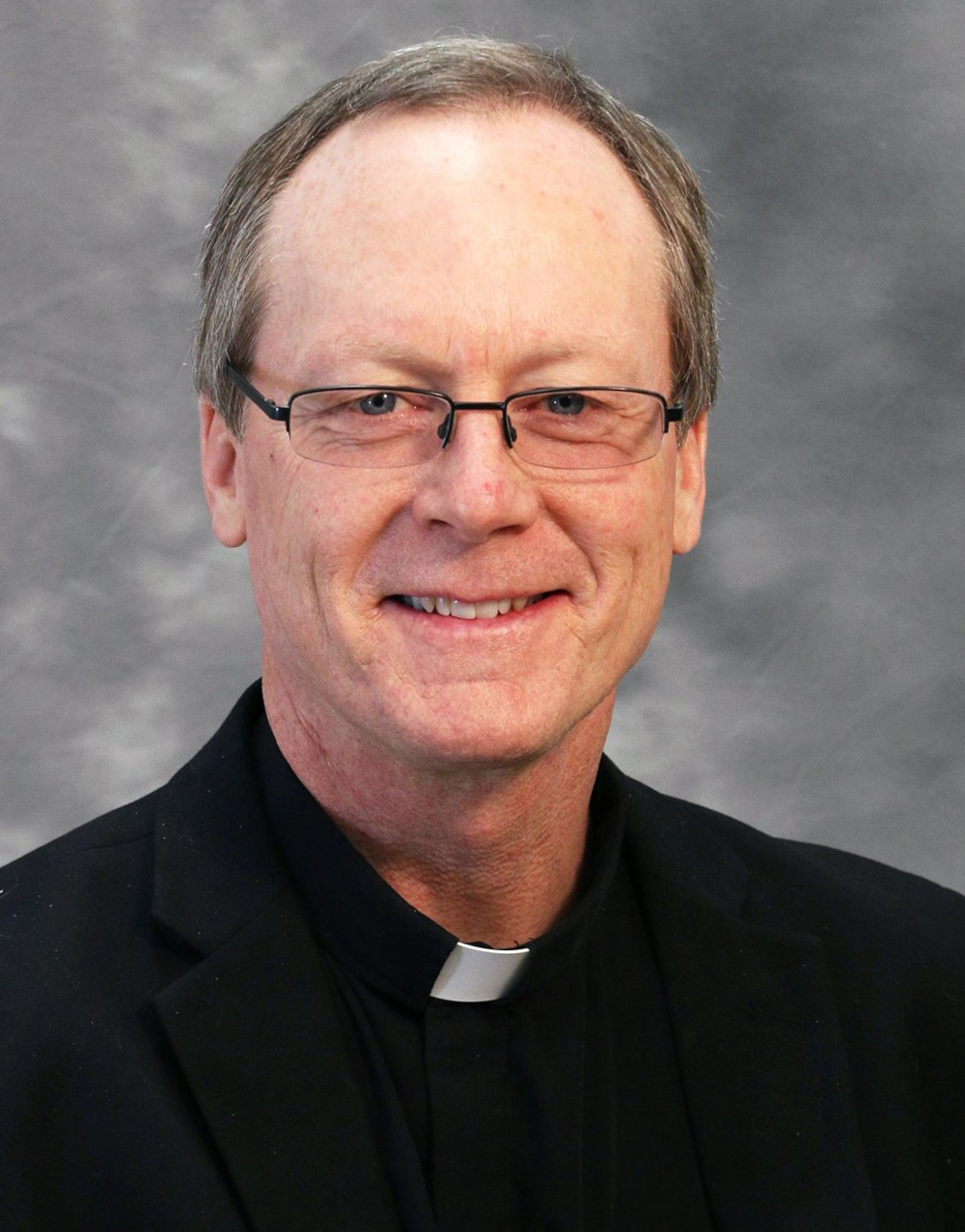 An older white man wearing glasses and a clerical collar smiles at the camera