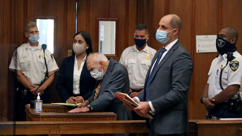 An older white man leans over behind a stand in a courtroom, where everyone is wearing masks