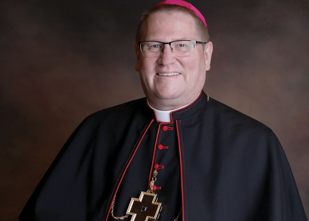 A white man wearing a violet zucchetto, glasses, and a bishop's cassock smiles at the camera