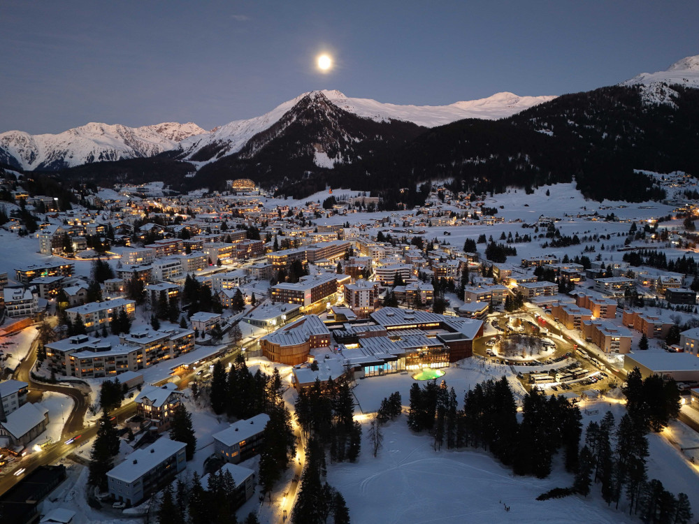 A snowy mountain town seen from above is lit up with warm lights below the full moon