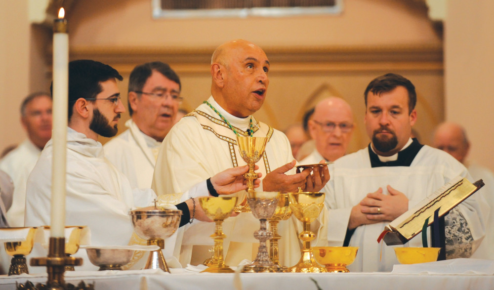 A bald man in white vestments holds a paten and speaks. Other priests in white surround him behind the altar.