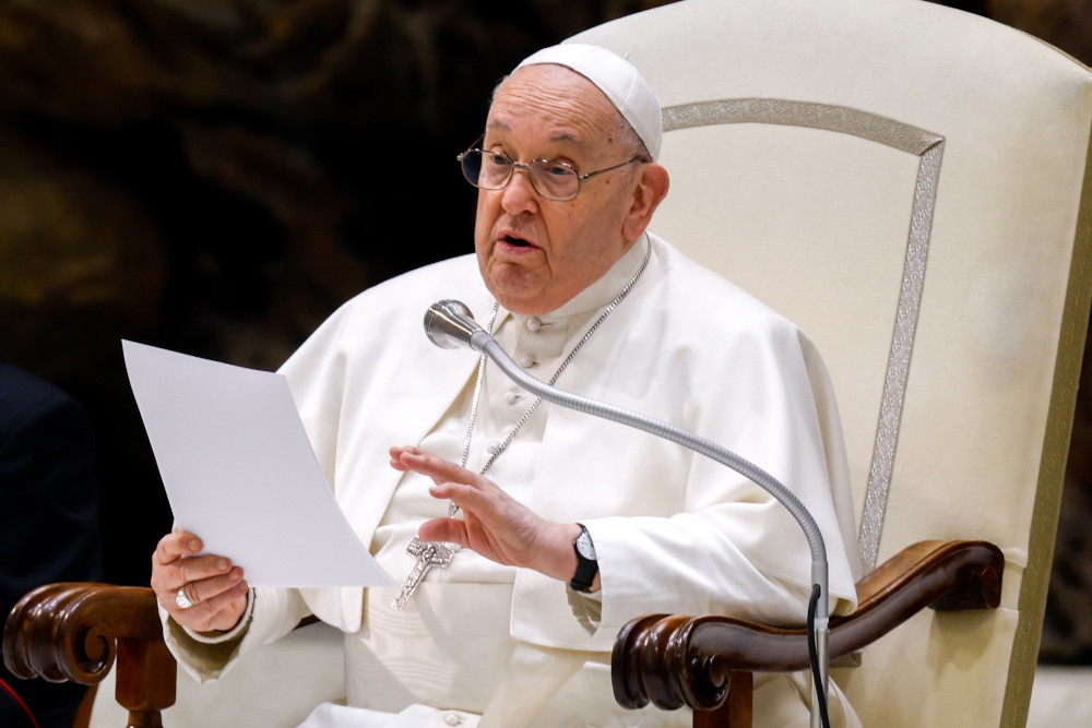 Pope Francis sits in his white chair and speaks into a microphone while holding a piece of paper