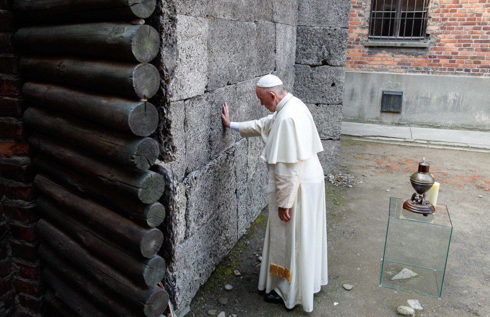 Pope Francis touches a gray wall while bowing his head. He stands in an outdoor space.
