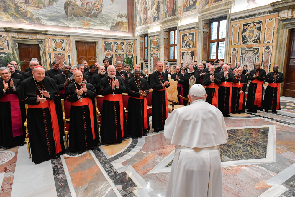Pope Francis faces and stands in front of a group of men wearing cardinals' cassocks