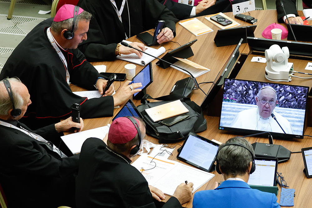 Participants watch Pope Francis on a video screen as they work on their tablets with synod documents during the first session of the assembly of the Synod of Bishops in the Paul VI Audience Hall at the Vatican Oct. 4, 2023. (CNS/Lola Gomez)