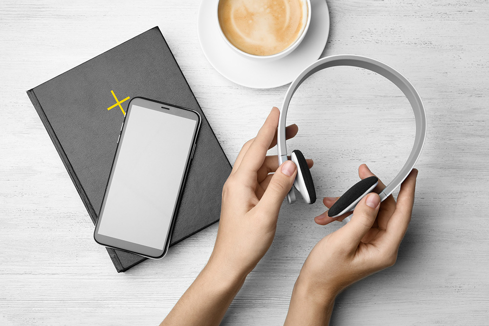 A woman's hands hold headphones next to a Bible with a smartphone on top and a cup of coffee (Dreamstime/Chernetskaya)