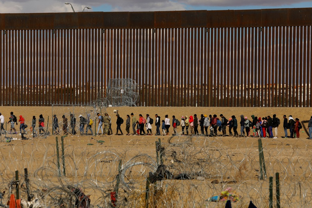 Asylum seekers border People seeking asylum lined before border wall in background.