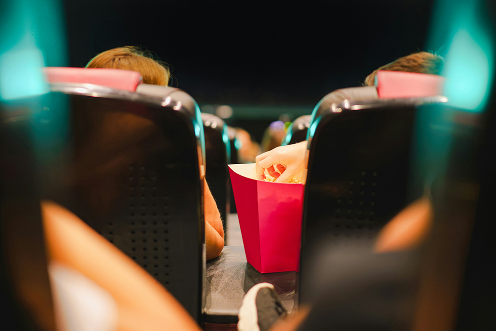 View of movie theater seats from behind, as one patron reaches for popcorn (Unsplash/Jakub Zerdzicki)