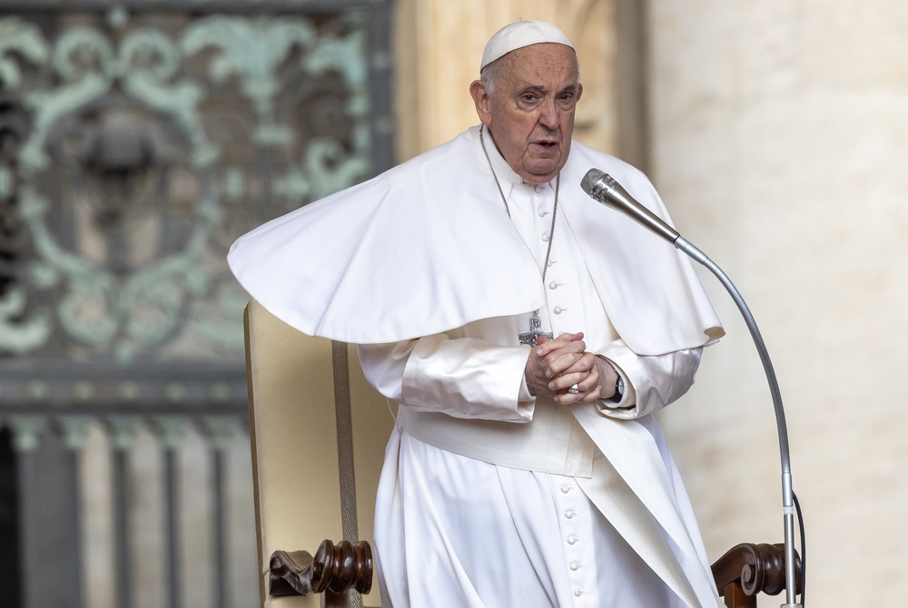 Pope Francis stands, hands folded, bracing against wind. 