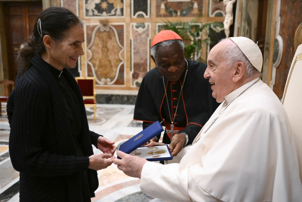 Pope Francis seated, accepts item from woman standing, cardinal bends to join.