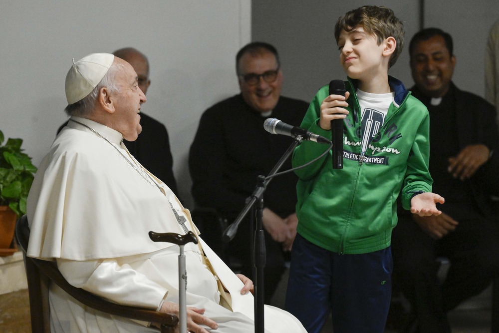 Child stands, with microphone, to ask Pope Francis, seated, a question; all smile. 