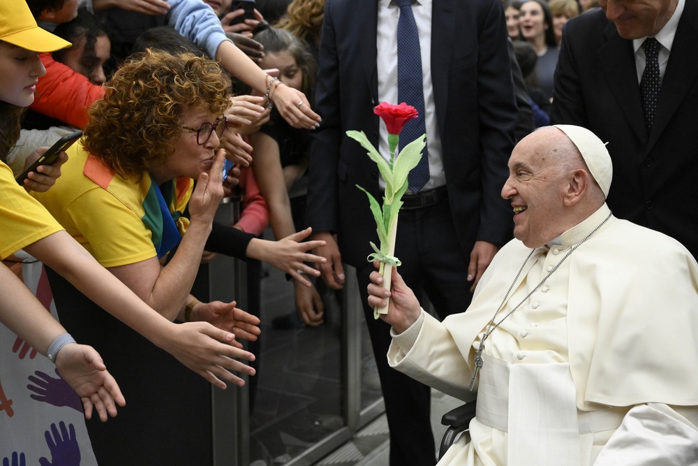 Pope Francis schools of peace Pope Francis, in wheelchair, holds flower, and smiles at woman who blows kiss from behind barrier.