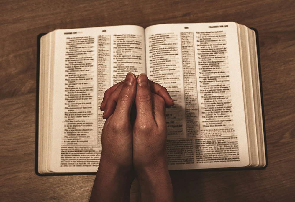 A reader holds folded hands on top of an open Bible that lays on a wooden surface. (Unsplash/Jametlene Reskp)