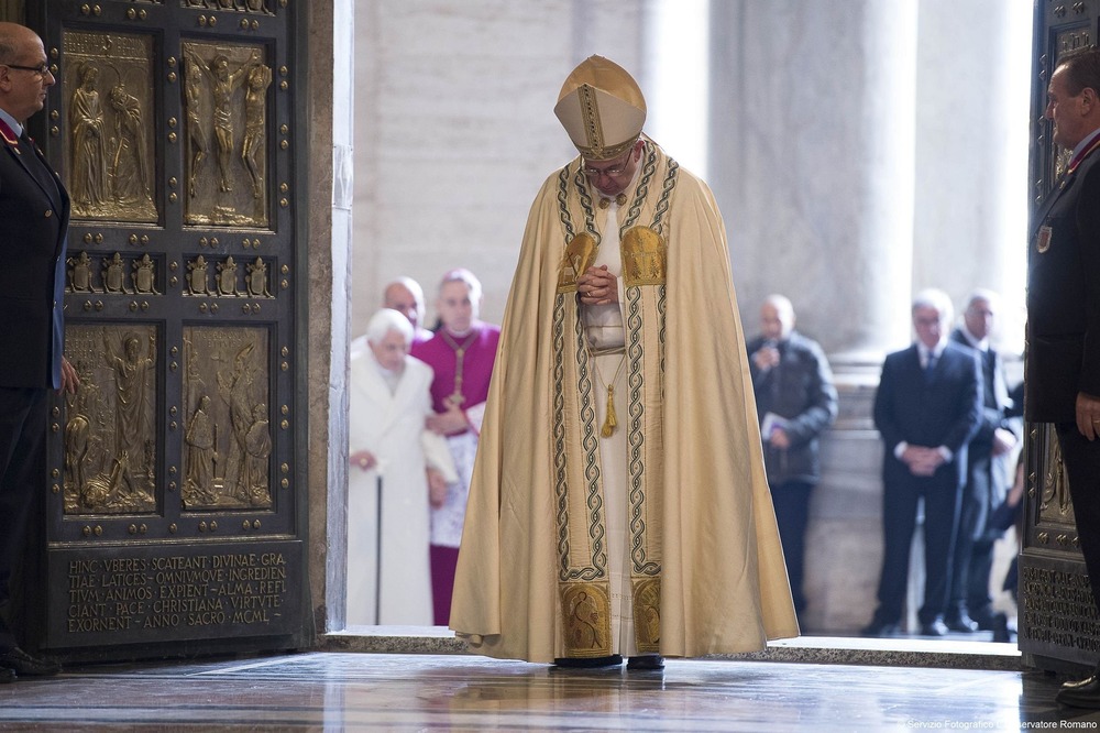 Pope Francis 2025 trip to Turkey Pope Francis, vested, walks with head bowed through Holy Doors of St. Peters; in background is Pope Emeritus Benedict XVI, wearing plain white coat, supported by cane and bishop.