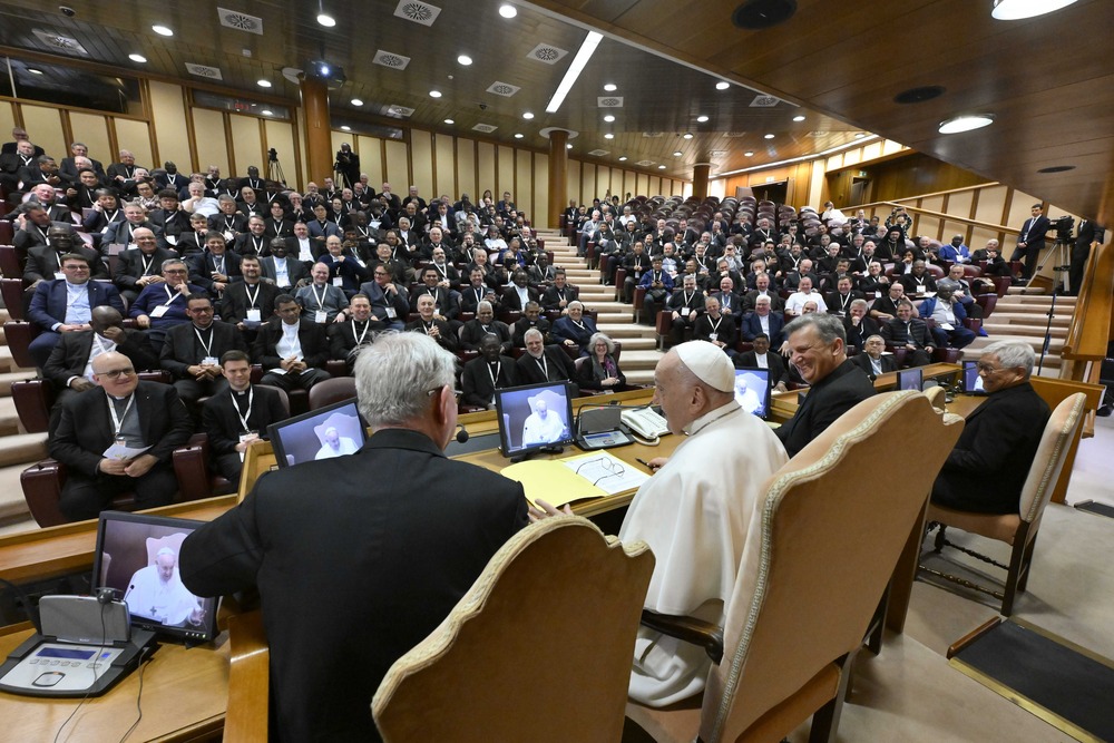 Pope Francis, synod pastors group. Pope pictured from behind, in front of him is audience hall filled with priests.