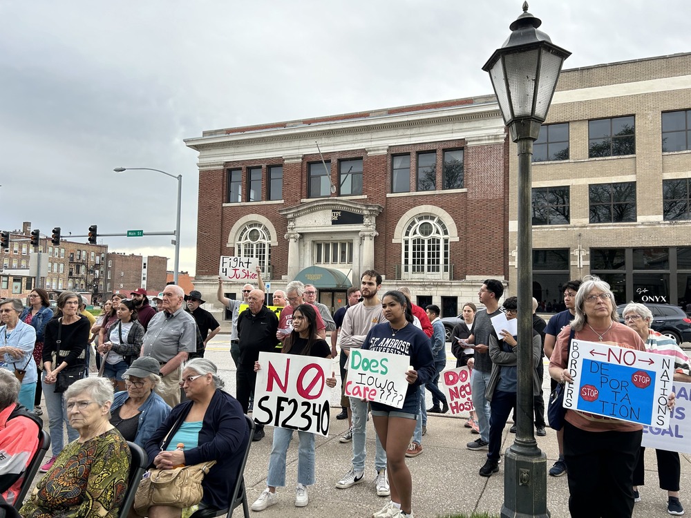 People holding signs demonstrate in square, with brick buildings in background. 