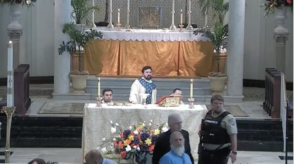 Abbeville, Louisiana Mass interrupted by armed teenager. Priest stands at altar while acolytes kneel and police officer scans crowd.