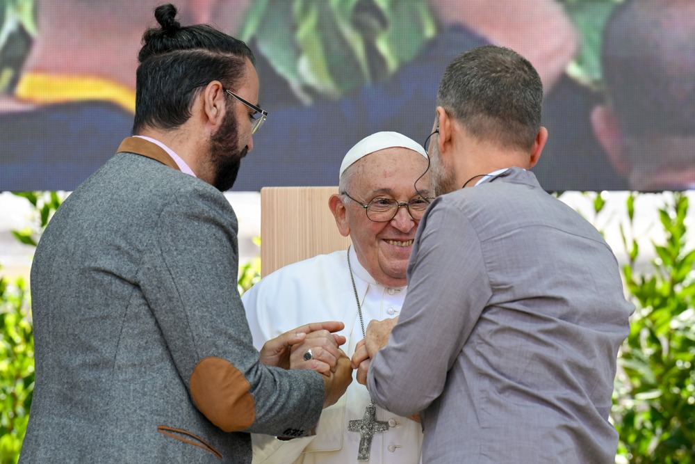 Pope Francis, Pentecost 2024 Vigil Mass, Verona, Italy. Pope Francis, seated, smiles and takes the hands of the two men.
