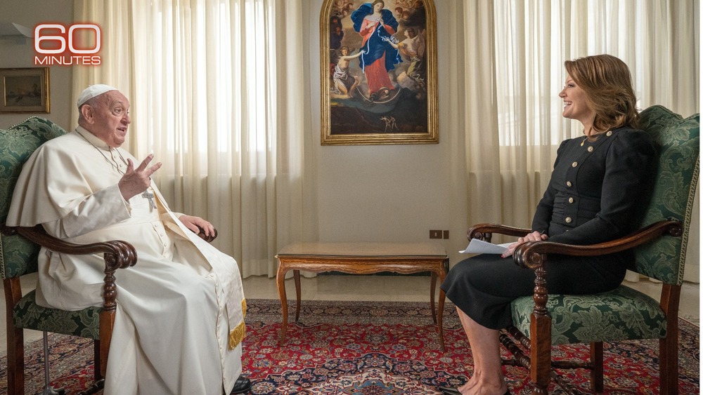Pope Francis 60 Minutes 2024 Pope Francis and Norah O'Donnell seated opposite one another, with painting and draped windows in background.