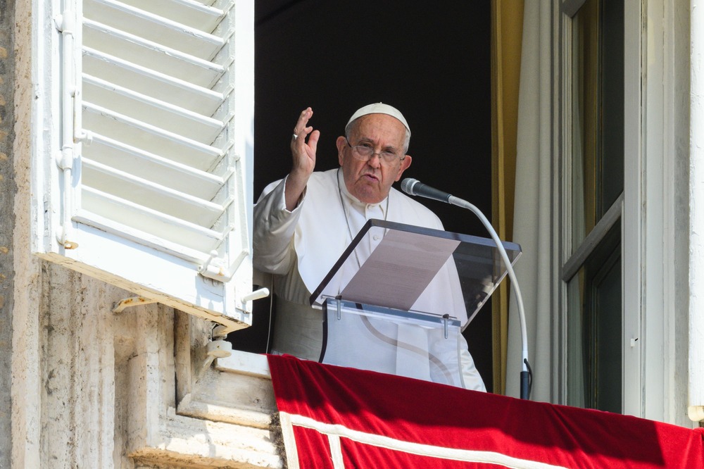 Pope Francis Angelus, July 21, 2024. Pope Francis raises hand in blessing, standing at a lectern on the balcony in St. Peter's Square.