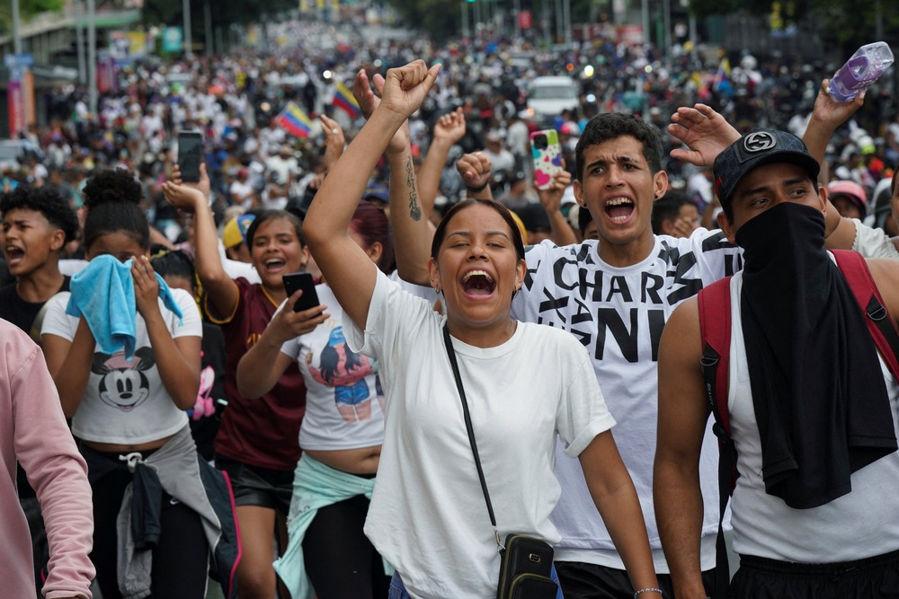 Opposition protest, Caracas, Venezuela, July 29, 2024. Woman in white shirt shouts chant while raising fist; behind hundreds of white-clothed people assemble and march.