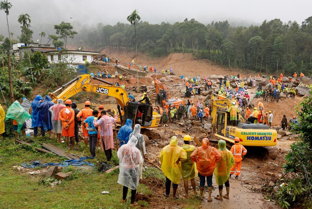 Landslide survivor search site in Kerala, India, 2024. Group of people wearing rain ponchos stand around search site along with earth movers.