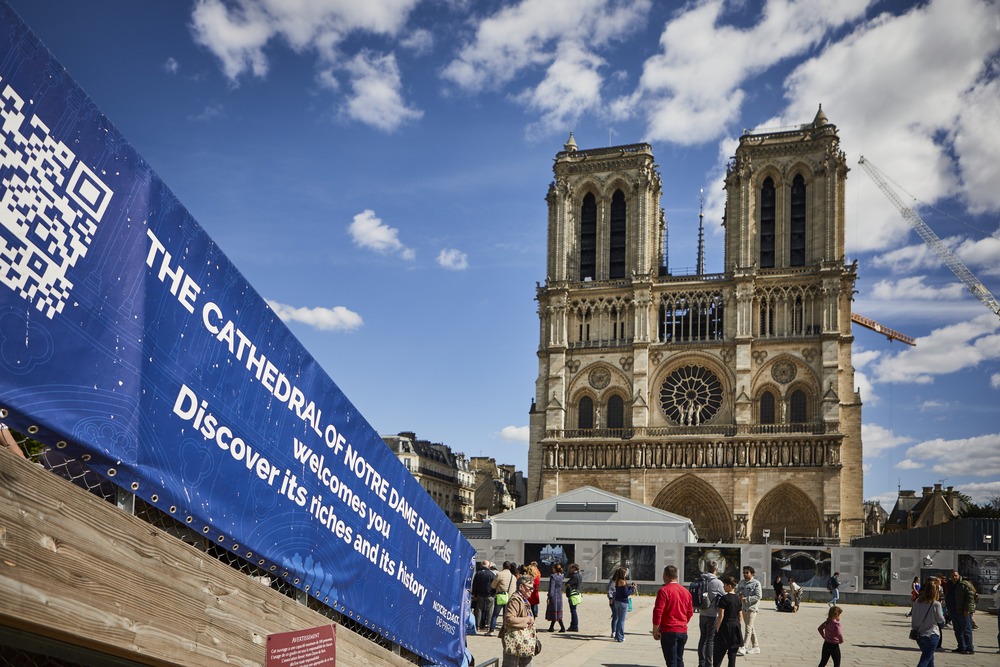 Notre Dame de Paris seen in background against bright blue sky; foregrounded is a tarp banner announcing the reconstruction and reopening of the Cathedral. 