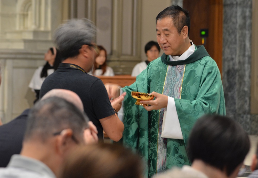 Bishop Paul Junmin Pei of Liaoning, China, in Chicago Aug.3, 2024. Pei, vested in green, hands Eucharistic host to person.