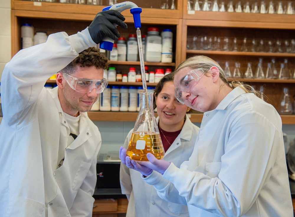 University of Mary SURVE students, Bismark, North Dakota, June 18, 2024. Three students wear white lab coats, and observe as one student uses a dropper to insert fluid into Erlenmeyer flask.