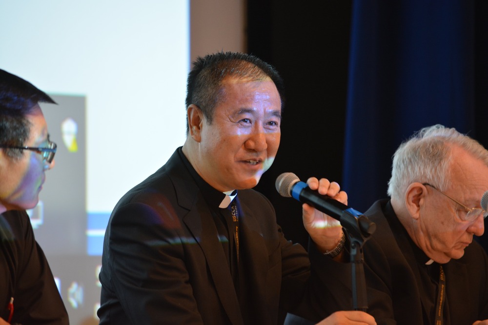 Bishop Paul Junmin Pei of Liaoning, China, in Chicago Aug.3, 2024. Bishop Pei sits at panel, wearing black clericals, and speaking into microphone.