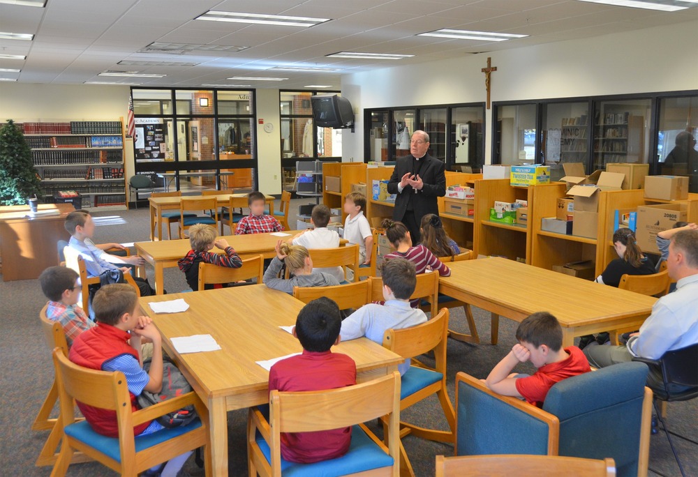 St. Dominic Academy, a K-12 school in the Diocese of Portland, Maine. Children sit at table in large room, while priest stands talking.