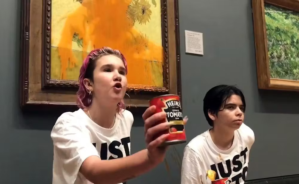 Just Stop Oil action, London 2023. Two young people wearing matching t-shirts, stand in front of painting; one holds can of soup, whose contents are splattered across painting.