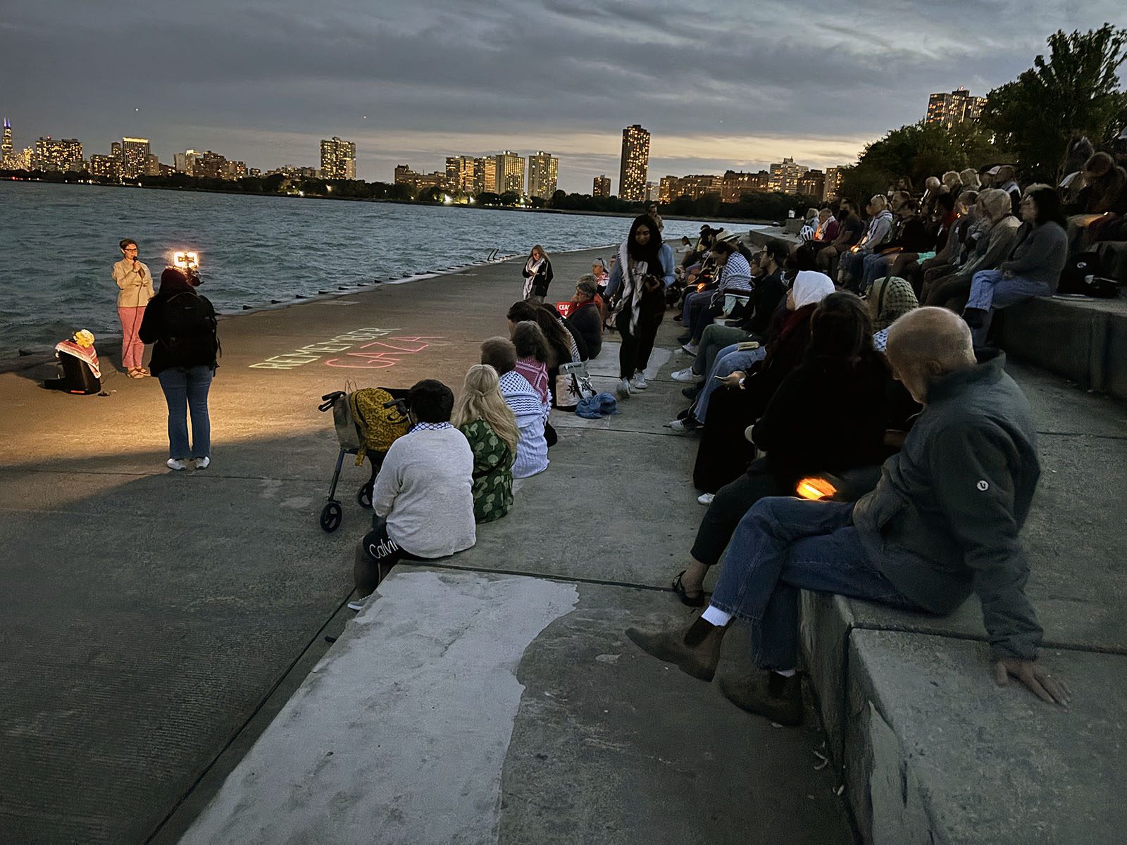 DNC Gaza protests, Aug. 20, 2024, Chicago. Chicago skyline at dusk in background; people sit gathered around speakers at edge of Lake Michigan.