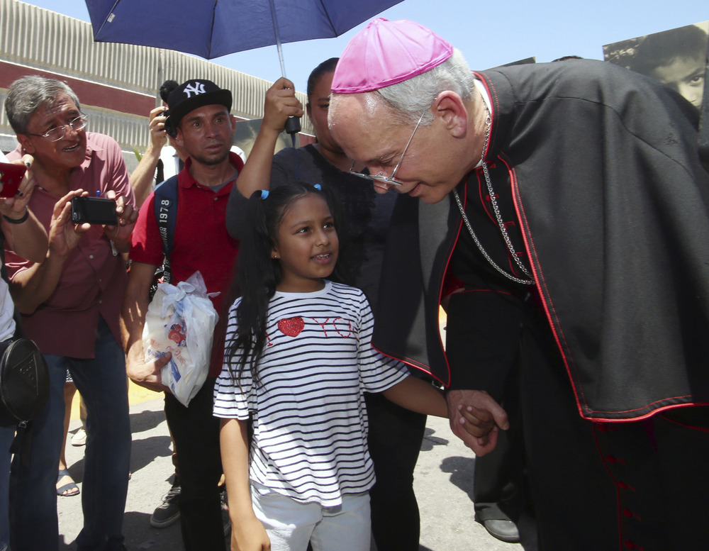 Bishop Mark Seitz of El Paso, Texas, and Celsia Palma, Juarez, Mexico, June 27, 2019. Seitz in purple-piped cassock and purple zucchetto, bends down to take little girl's hand.
