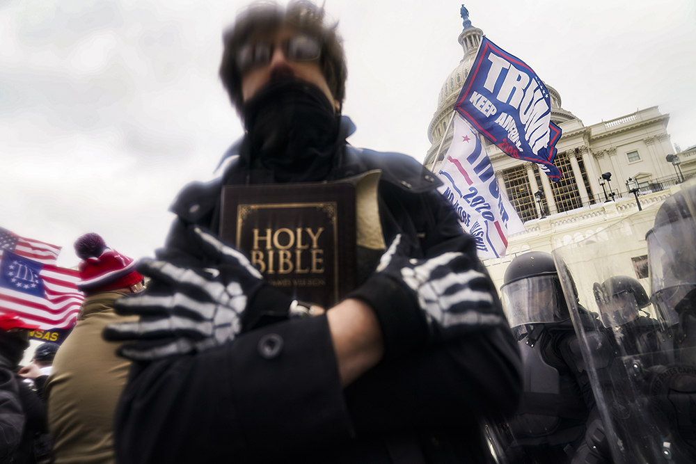 Jan. 6, 2021 Capitol riot, Washington D.C.. Rioter with face obscured, holds bible to chest, in the background is Capitol building dome and various flags, including one bearing former president Donald Trump's last name.
