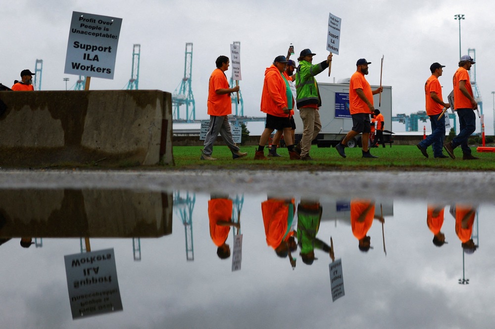 International Longshoresmen's Association members picketing at the Virginia International Gateway in Portsmouth, Oct. 1, 2024. Picket line viewed from side, as workers wearing reflective orange gear and holding signs walk by; they are reflected in a pool of water that occupies the bottom half of the frame.