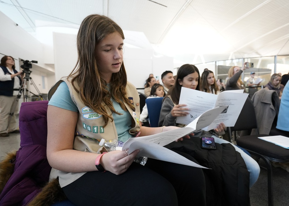 Girl Scouts, New York, Dec. 4, 2022. Girl wearing scout vest and holding program fills frame. Behind are other troopers singing.