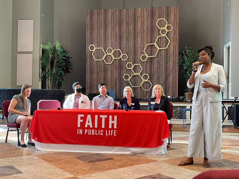 Jeanné Lewis, Faith in Public Life, Georgia, Aug. 29, 2024. Lewis stands speaking into mic in front of panel with five other people seated.
