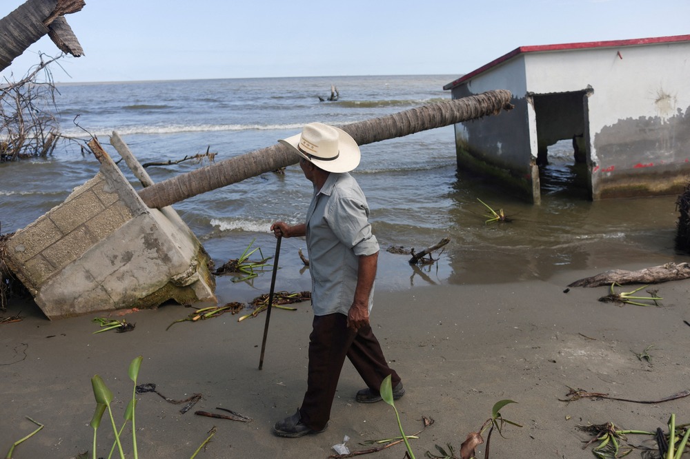 Rising sea-level damage, El Bosque, Mexico, Nov. 7, 2022. Man with walking stick foregrounded, in background are building ruins and the shoreline.