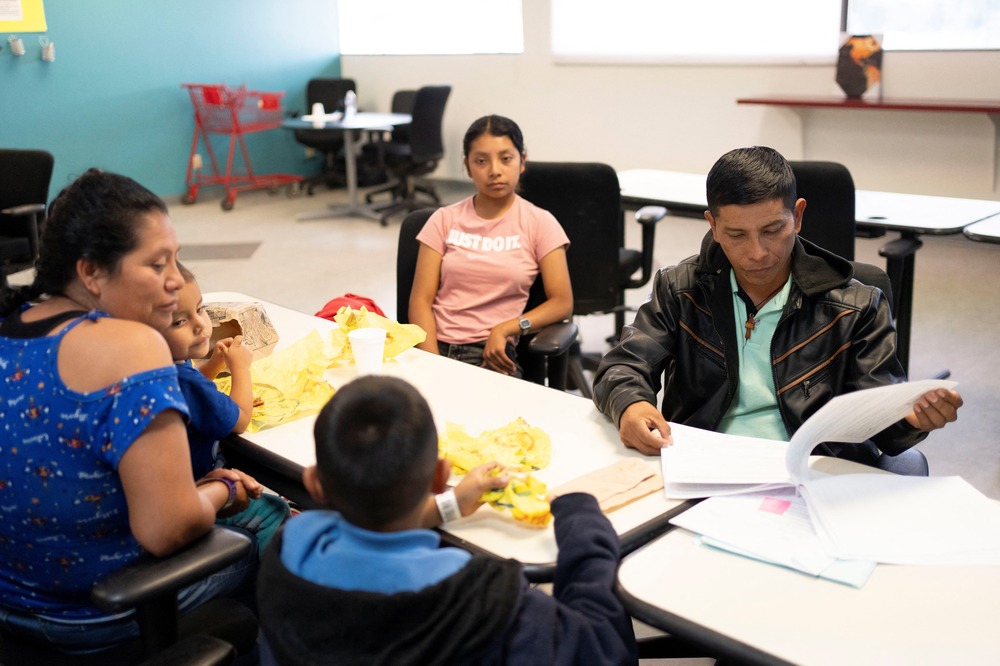 Migrant family from Chiapas, Mexico, Tucson, Arizona, March 15, 2024. Family sits around table with trays of food in what looks like a classroom.