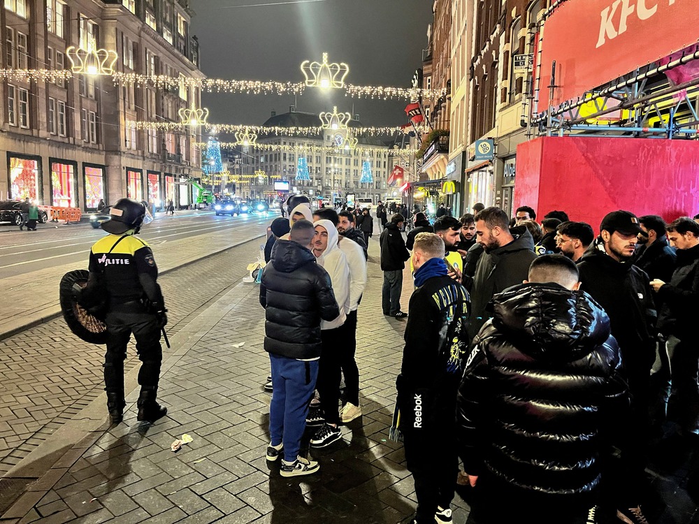 Israeli soccer fans protected by police following riots, Amsterdam, Nov. 8, 2024. People stand on street wearing coats, assembled are police.